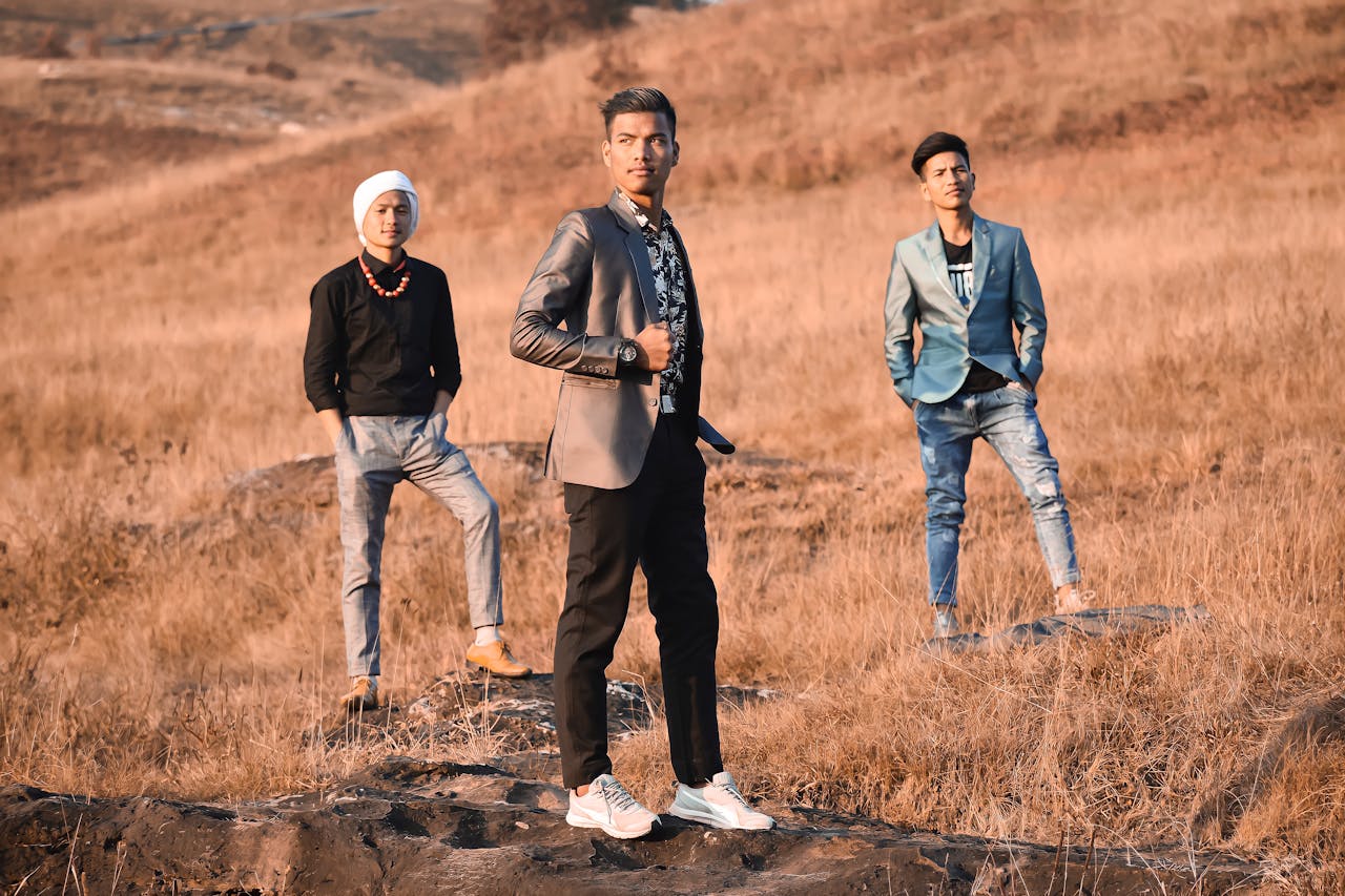 Three young men in fashionable attire posing stylishly in an open field during the day.