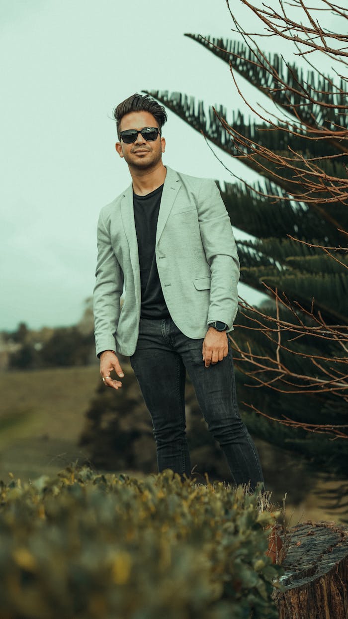 Confident man in blazer and sunglasses, posing outdoors with a smile.